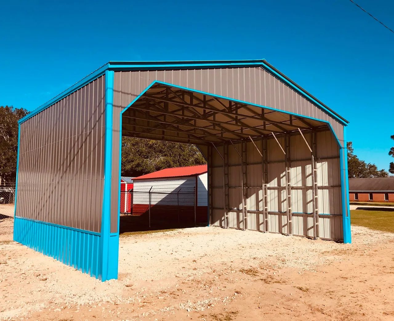 Vertical-roof metal carport with blue trim and enclosed sides, open at the front, on a sand pad