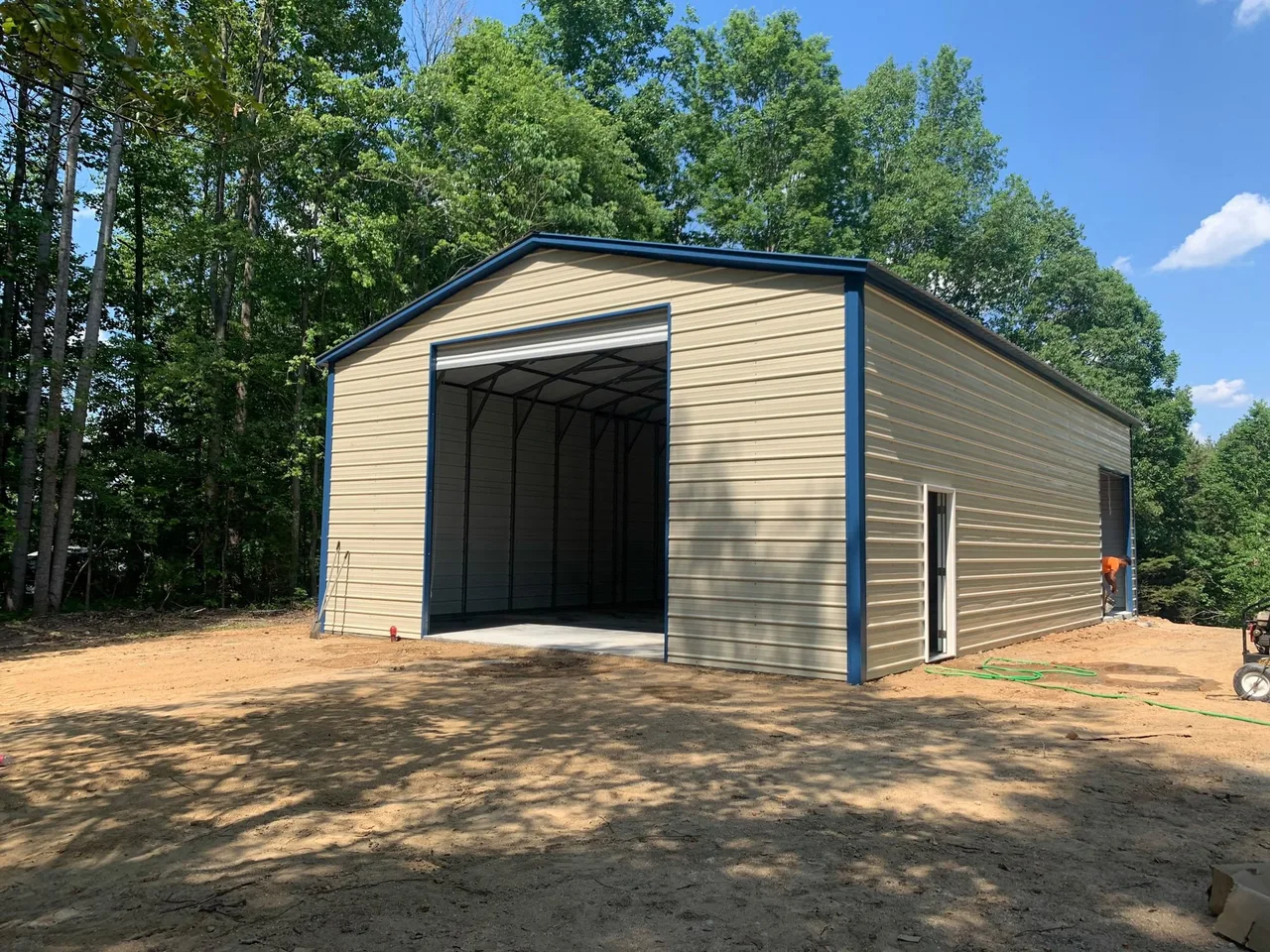 Tall tan metal workshop with blue trim and a large open roll-up door, among trees