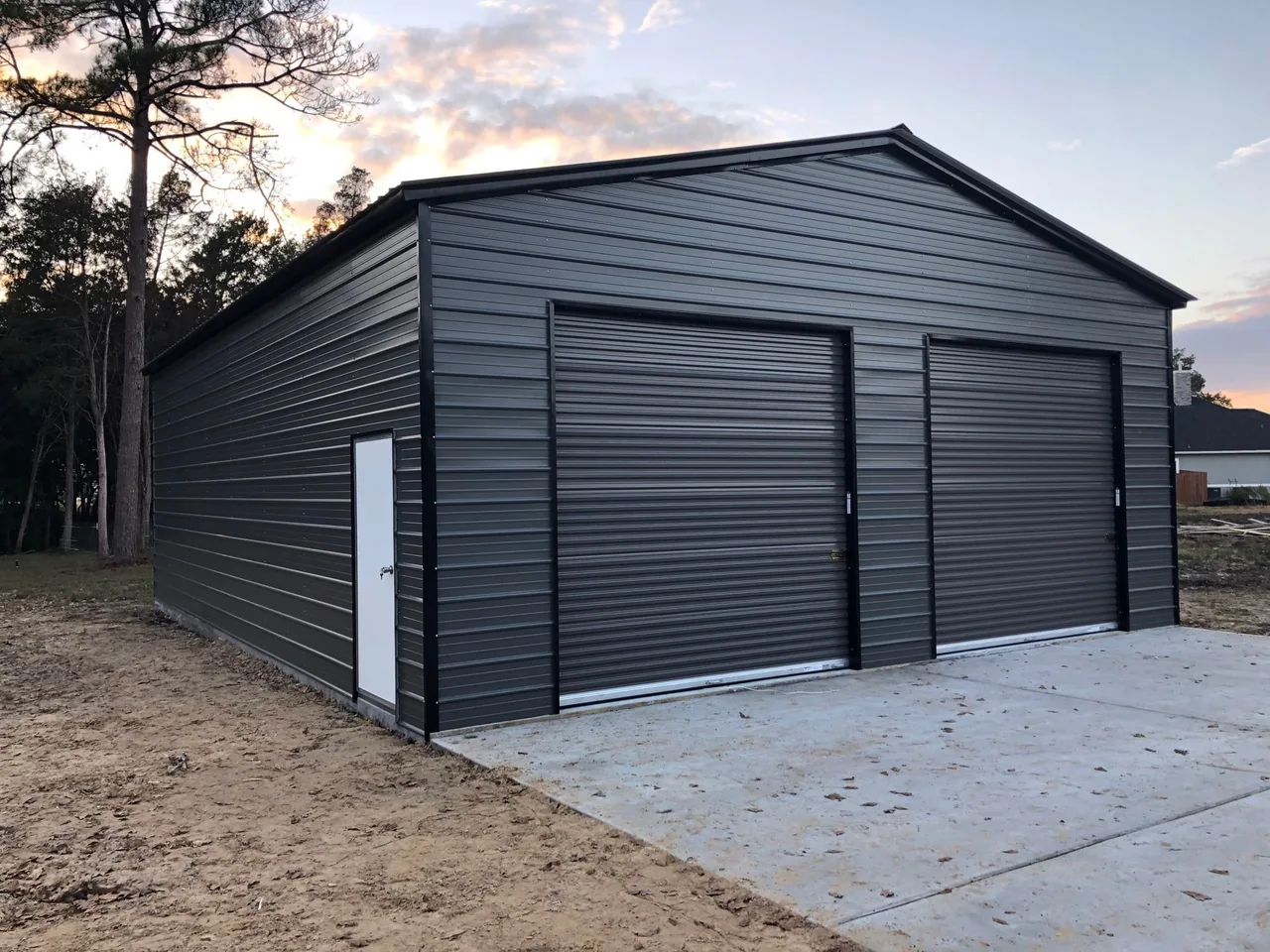Dark gray two-car metal garage with two roll-up doors and a walk-in door, on a new concrete slab